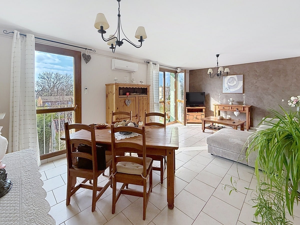 Séjour rectangulaire avec parquet au sol et murs blancs. Baie vitrée coulissante ouvrant sur un balcon avec garde-corps métallique, stores vénitiens aux fenêtres.