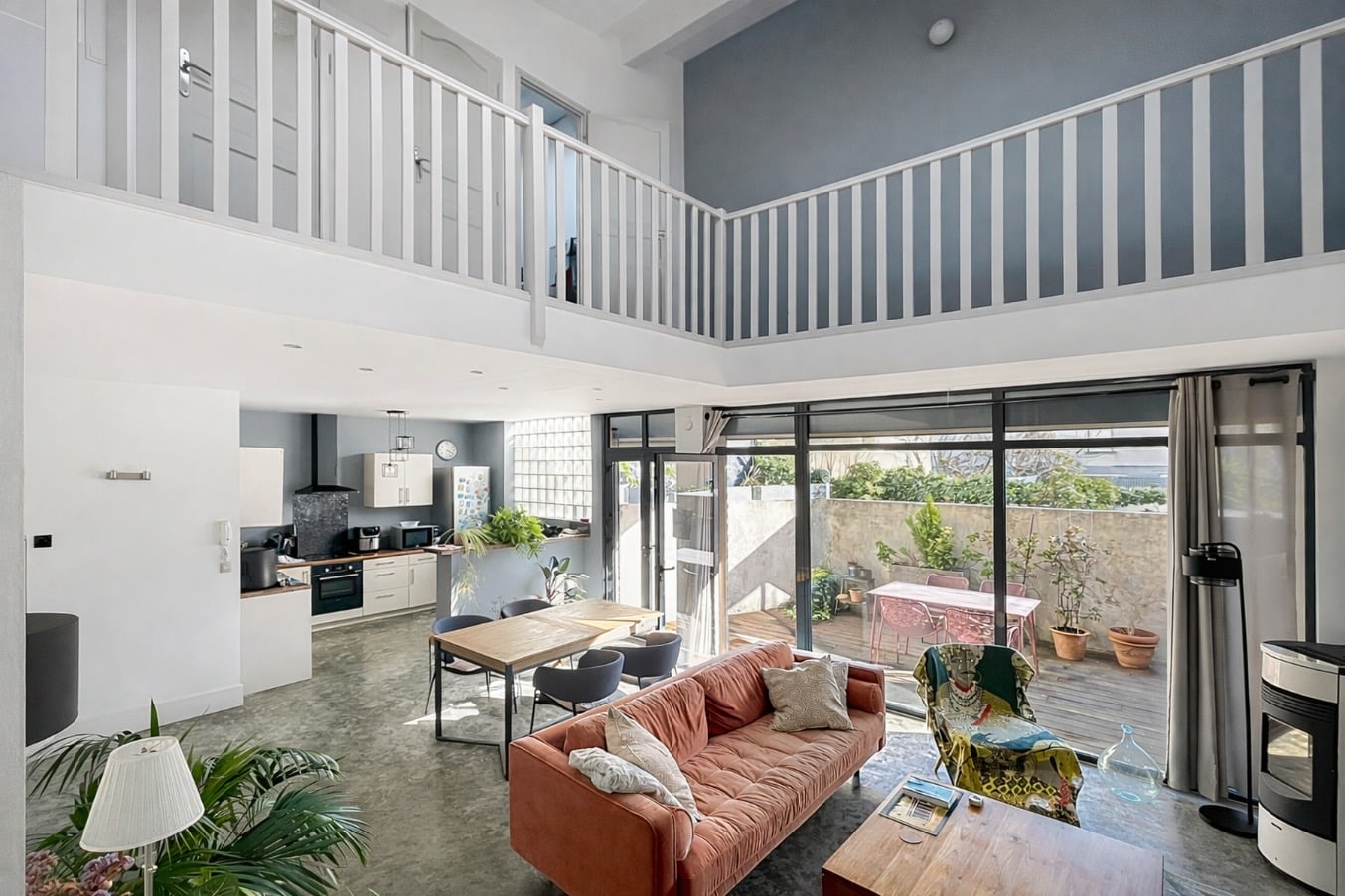 Pièce de vie à volume double hauteur avec mezzanine et garde-corps blanc, cuisine ouverte visible en arrière-plan et terrasse accessible par baies vitrées. Vue d'ensemble montrant la hauteur sous plafond, le poêle à granulés et le sol en béton ciré.