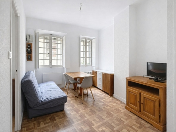 Pièce de vie meublée avec canapé, table à manger, meuble bas et télévision sur meuble en bois. Sol en parquet mosaïque, murs blancs et deux fenêtres à petits carreaux en bois caractéristiques du bâti ancien.