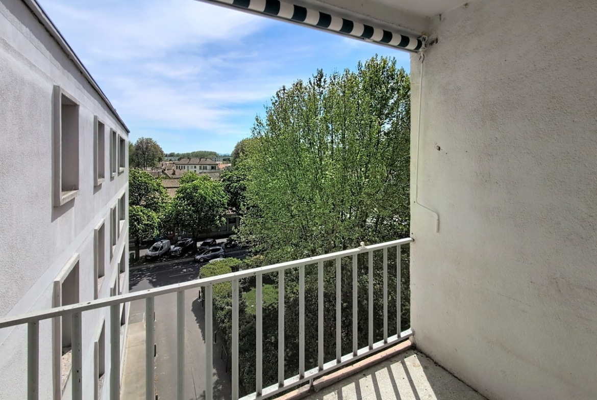 Balcon avec garde-corps à barreaux blancs et store banne rayé, vue sur parking arboré et toits de la ville. Ciel bleu avec quelques nuages, façade crépi blanc visible.