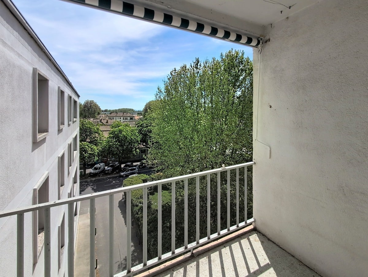 Balcon avec garde-corps à barreaux blancs et store banne rayé, vue sur parking arboré et toits de la ville. Ciel bleu avec quelques nuages, façade crépi blanc visible.