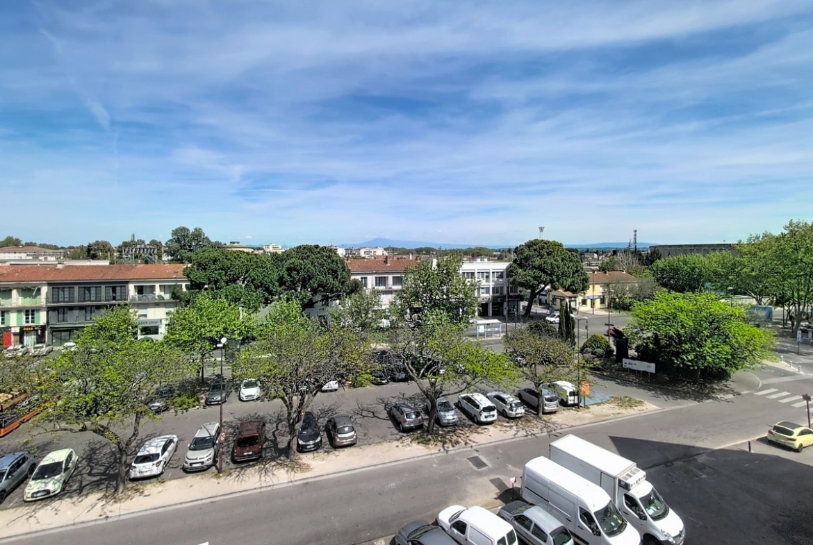 Vue aérienne depuis un étage élevé sur place de parking bordée d'arbres et commerces. Panorama dégagé vers les collines et silhouette du Mont Ventoux en arrière-plan.