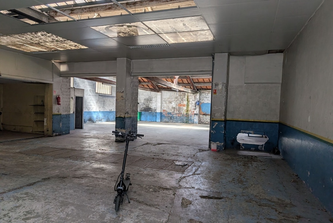 Espace intérieur de type atelier avec sol béton brut et trottinette électrique au centre. Plafond suspendu avec panneaux translucides laissant passer la lumière naturelle, murs peints en bleu et blanc avec traces d'usure, piliers en béton gris et extincteur rouge visible sur la gauche.