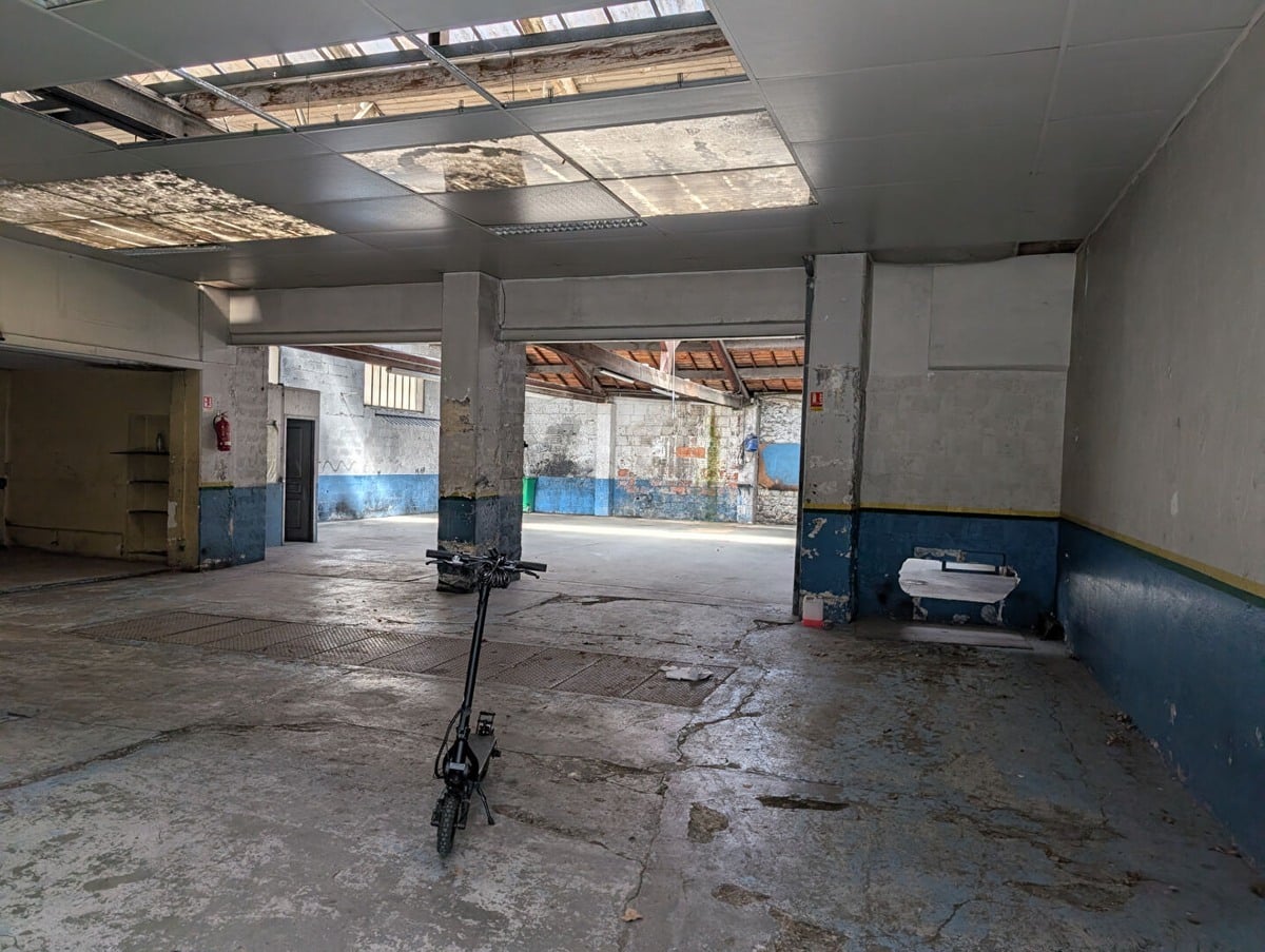 Espace intérieur de type atelier avec sol béton brut et trottinette électrique au centre. Plafond suspendu avec panneaux translucides laissant passer la lumière naturelle, murs peints en bleu et blanc avec traces d'usure, piliers en béton gris et extincteur rouge visible sur la gauche.
