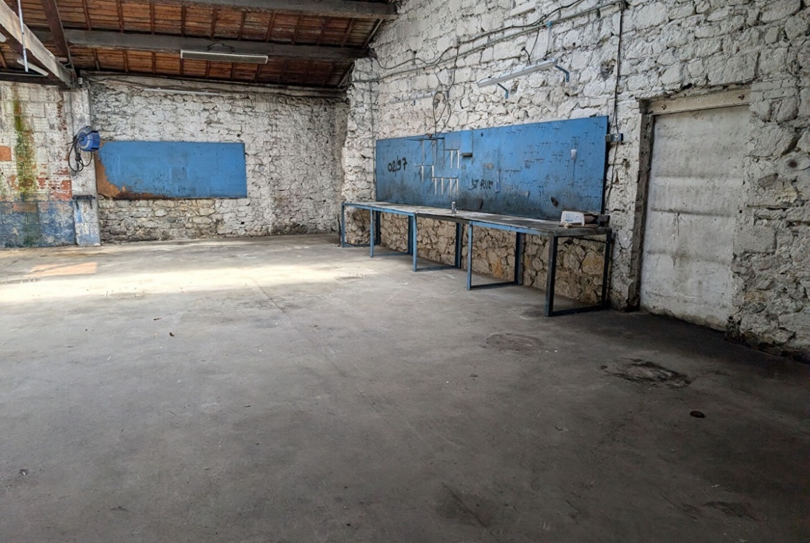 Espace de travail avec longs plans de travail métalliques bleus fixés contre des murs en pierre blanche apparente. Panneaux perforés bleus au-dessus des établis pour accrochage d'outils, toiture en tuiles avec charpente bois visible, sol béton présentant des traces d'usage, câbles électriques apparents.