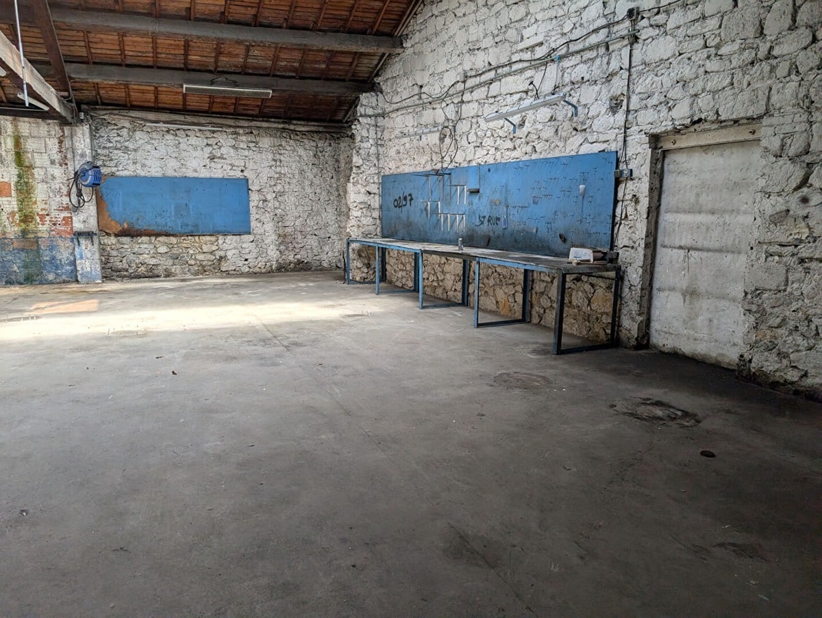 Espace de travail avec longs plans de travail métalliques bleus fixés contre des murs en pierre blanche apparente. Panneaux perforés bleus au-dessus des établis pour accrochage d'outils, toiture en tuiles avec charpente bois visible, sol béton présentant des traces d'usage, câbles électriques apparents.