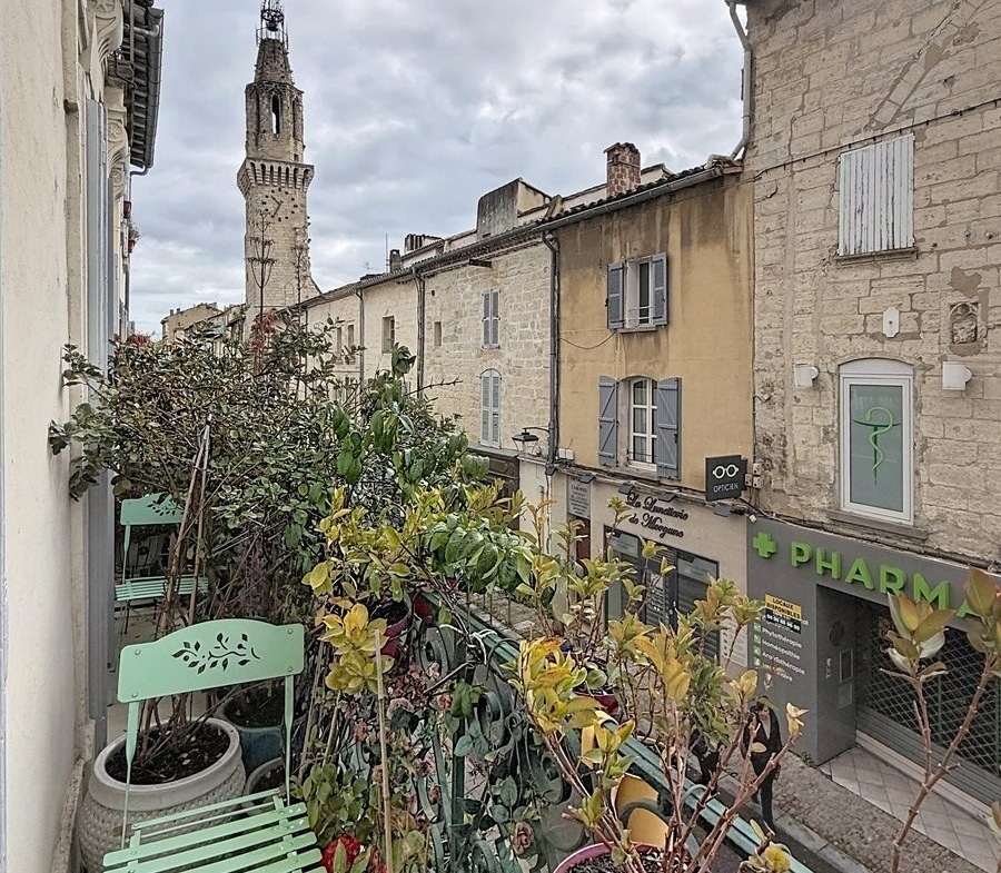 Balcon fleuri avec jardinières contenant plantes vertes et fleurs rouges, chaise pliante vert menthe. Vue sur rue pavée du centre historique avec façades en pierre, pharmacie en rez-de-chaussée et clocher médiéval ajouré en arrière-plan sous ciel nuageux.