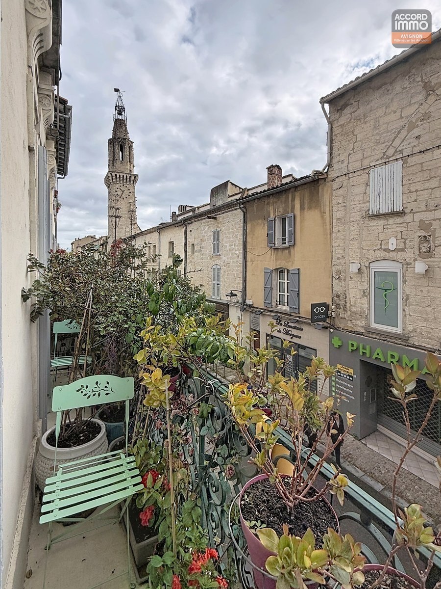 Balcon fleuri avec jardinières contenant plantes vertes et fleurs rouges, chaise pliante vert menthe. Vue sur rue pavée du centre historique avec façades en pierre, pharmacie en rez-de-chaussée et clocher médiéval ajouré en arrière-plan sous ciel nuageux.
