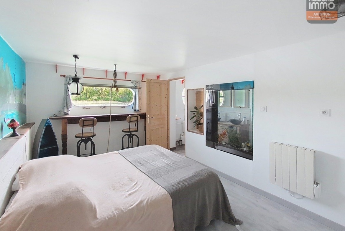 Chambre avec lit double recouvert d'un plaid gris, équipée d'un coin bar avec deux tabourets industriels face à une fenêtre panoramique. Mur d'accent turquoise à gauche, porte en bois clair menant à une salle d'eau visible à travers une ouverture vitrée, radiateur blanc et sol en parquet clair.