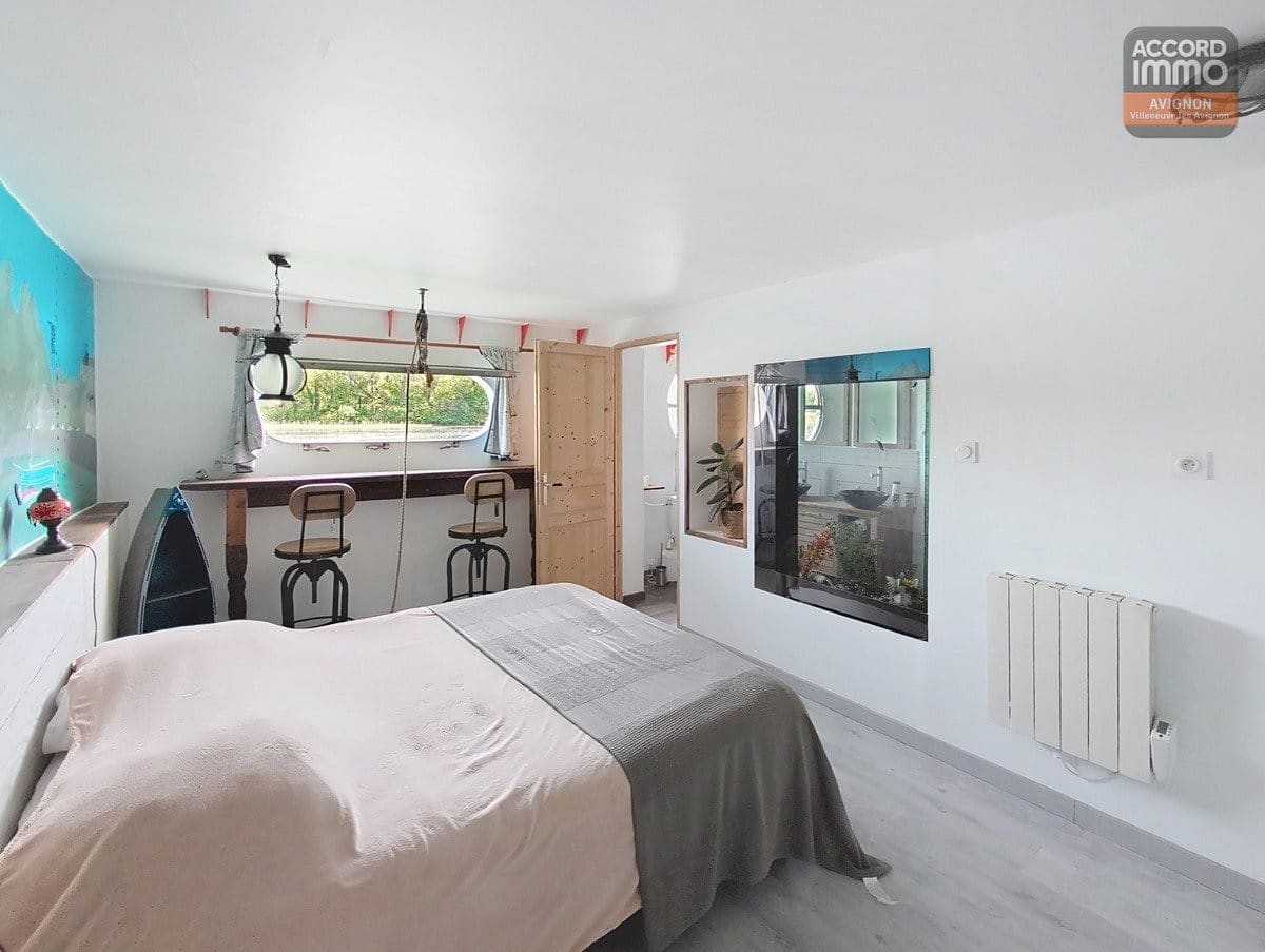 Chambre avec lit double recouvert d'un plaid gris, équipée d'un coin bar avec deux tabourets industriels face à une fenêtre panoramique. Mur d'accent turquoise à gauche, porte en bois clair menant à une salle d'eau visible à travers une ouverture vitrée, radiateur blanc et sol en parquet clair.