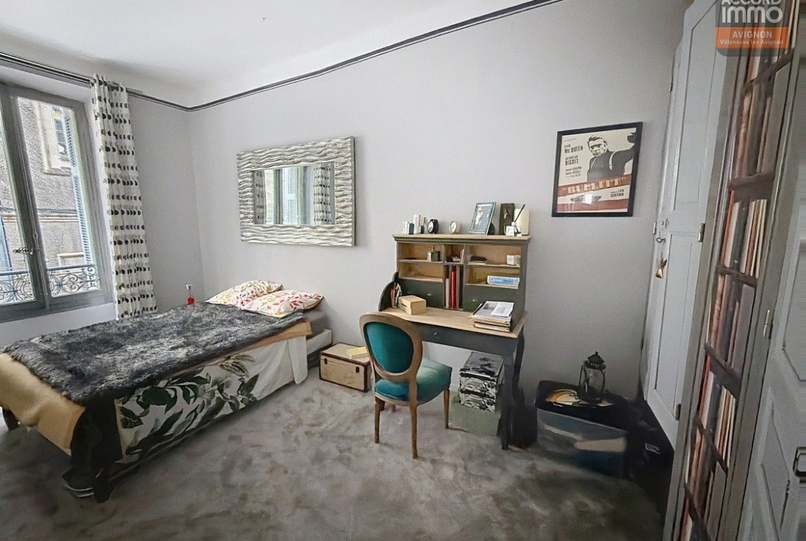 Chambre avec lit simple recouvert d'un plaid gris et coussins colorés, bureau secrétaire en bois naturel avec chaise velours turquoise. Murs gris clair, miroir mural ondulé, poster de film Steve McQueen Bullitt, sol en pierre naturelle beige et fenêtre avec balcon en fer forgé.