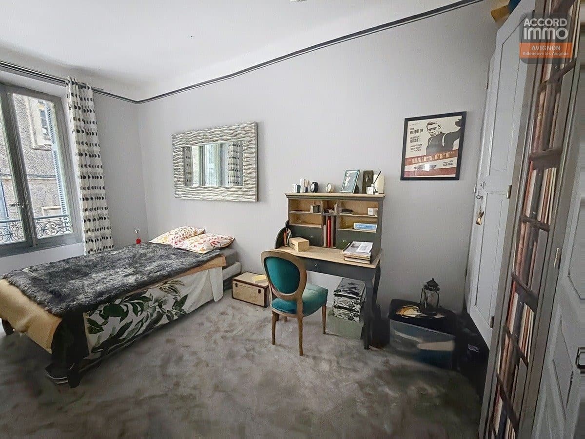 Chambre avec lit simple recouvert d'un plaid gris et coussins colorés, bureau secrétaire en bois naturel avec chaise velours turquoise. Murs gris clair, miroir mural ondulé, poster de film Steve McQueen Bullitt, sol en pierre naturelle beige et fenêtre avec balcon en fer forgé.