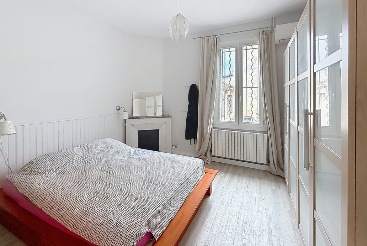 Chambre avec lit double à cadre bois et linge de lit à carreaux. Cheminée décorative blanche, armoire à portes vitrées et rideaux beiges encadrant une fenêtre avec grille.