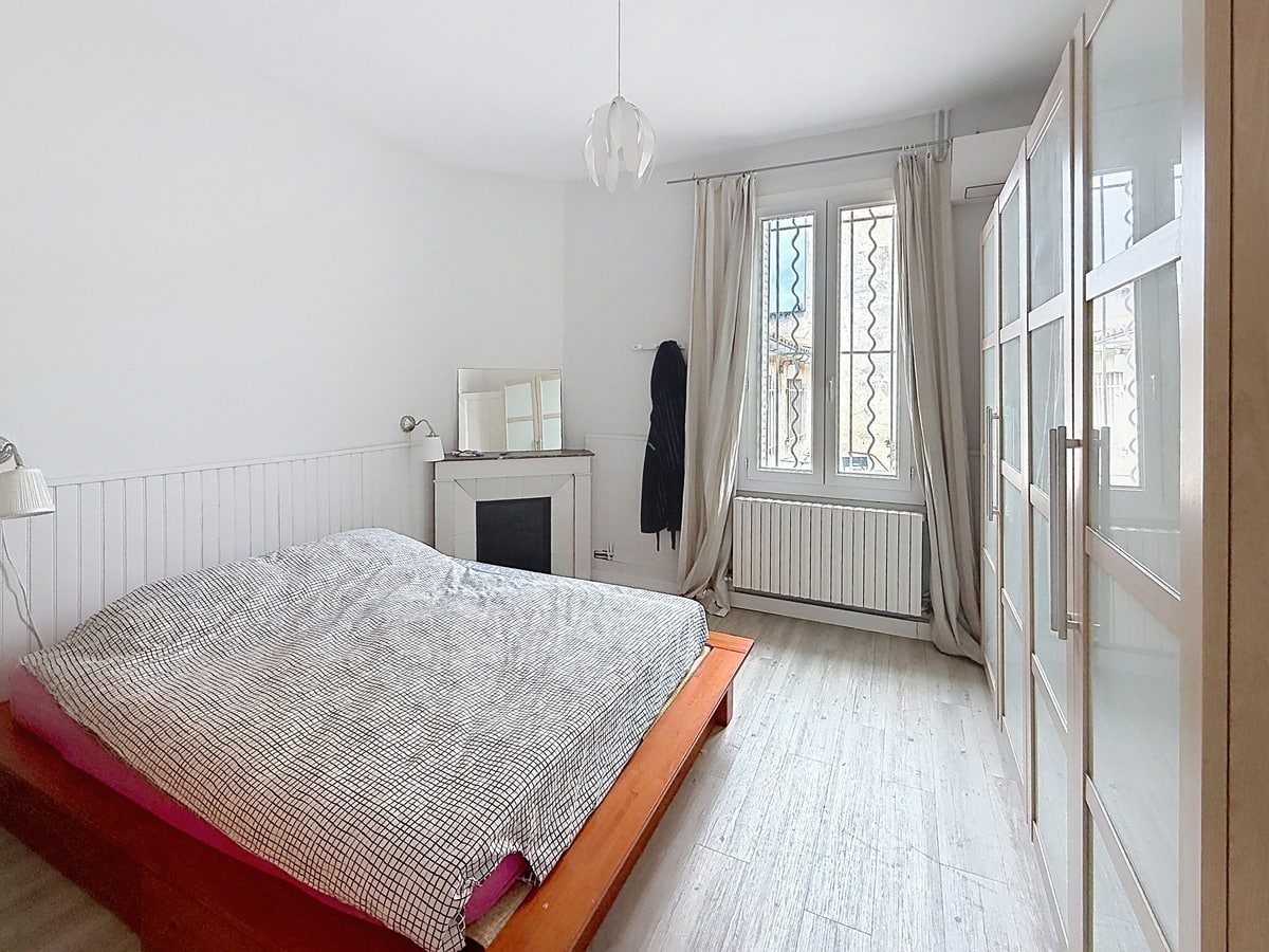 Chambre avec lit double à cadre bois et linge de lit à carreaux. Cheminée décorative blanche, armoire à portes vitrées et rideaux beiges encadrant une fenêtre avec grille.
