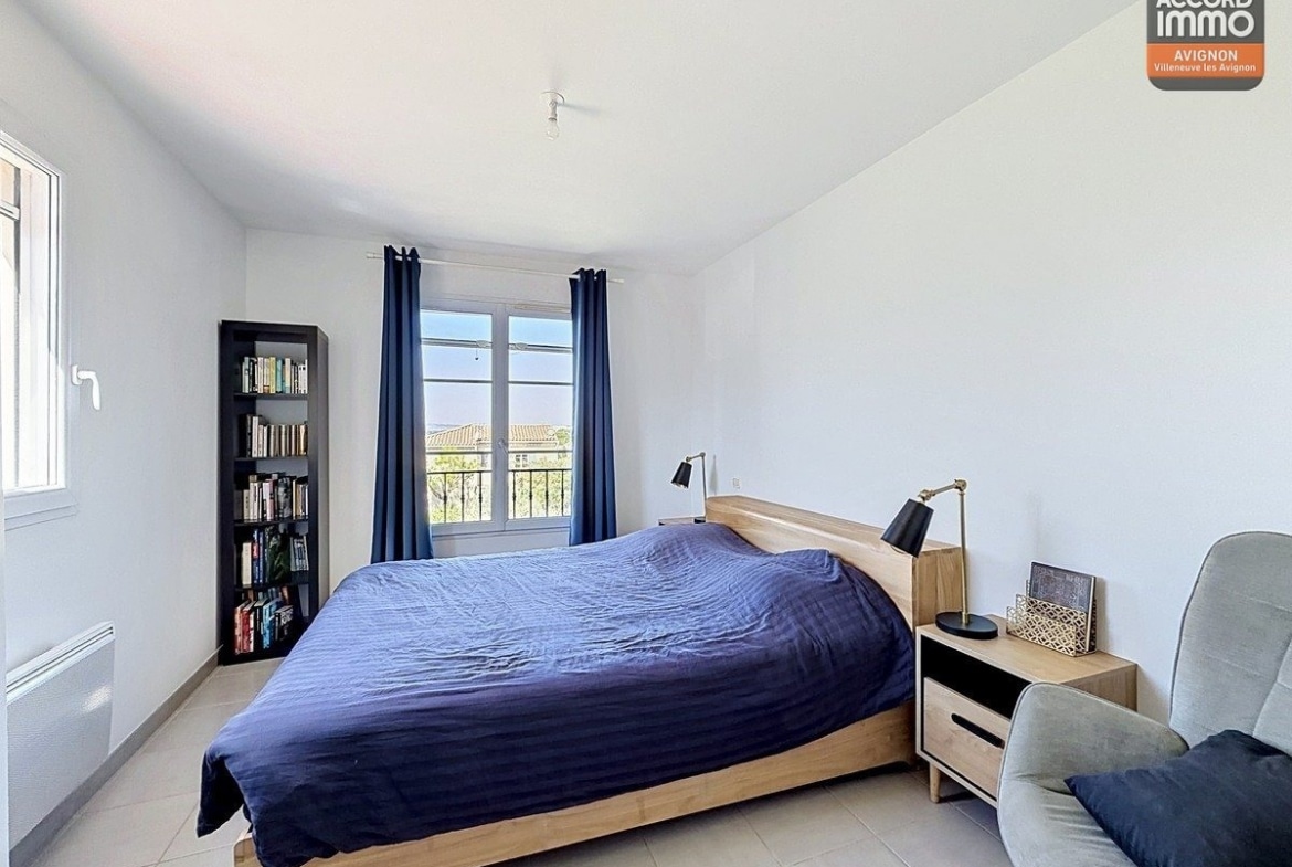 Chambre avec lit double recouvert d'une couette bleue et tête de lit en bois clair. Une bibliothèque noire remplie de livres se trouve près de la fenêtre avec rideaux bleus, et un fauteuil gris complète l'espace.
