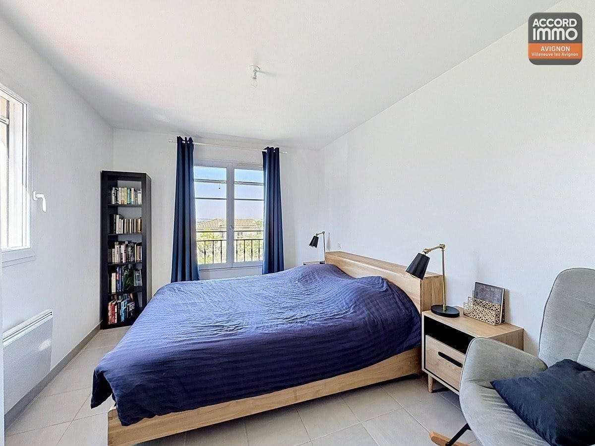 Chambre avec lit double recouvert d'une couette bleue et tête de lit en bois clair. Une bibliothèque noire remplie de livres se trouve près de la fenêtre avec rideaux bleus, et un fauteuil gris complète l'espace.
