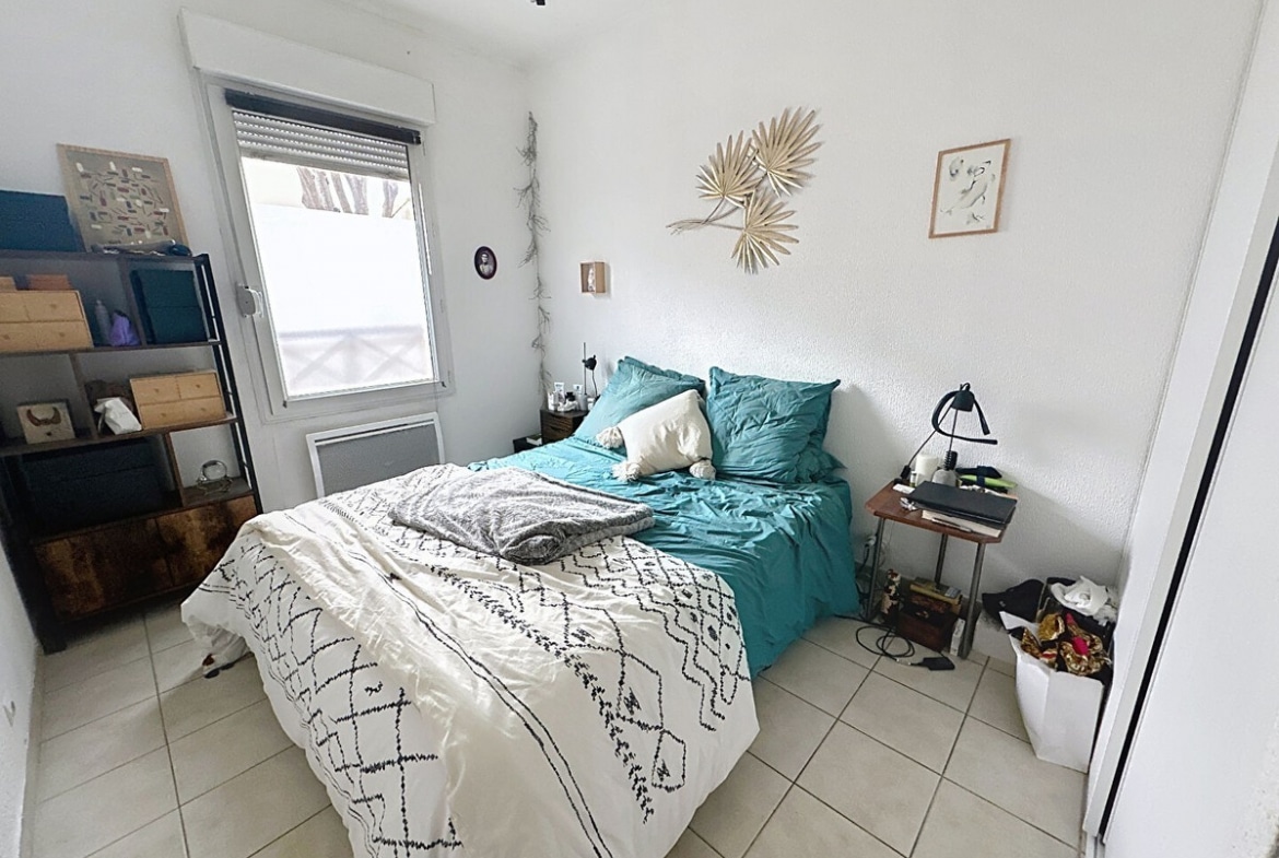 Chambre avec lit deux places, couette blanche à motifs géométriques noirs et oreillers turquoise. Étagère métallique noire avec boîtes de rangement, tables de chevet en bois de part et d'autre, décoration murale en feuilles de palmier séchées et fenêtre avec volet roulant.