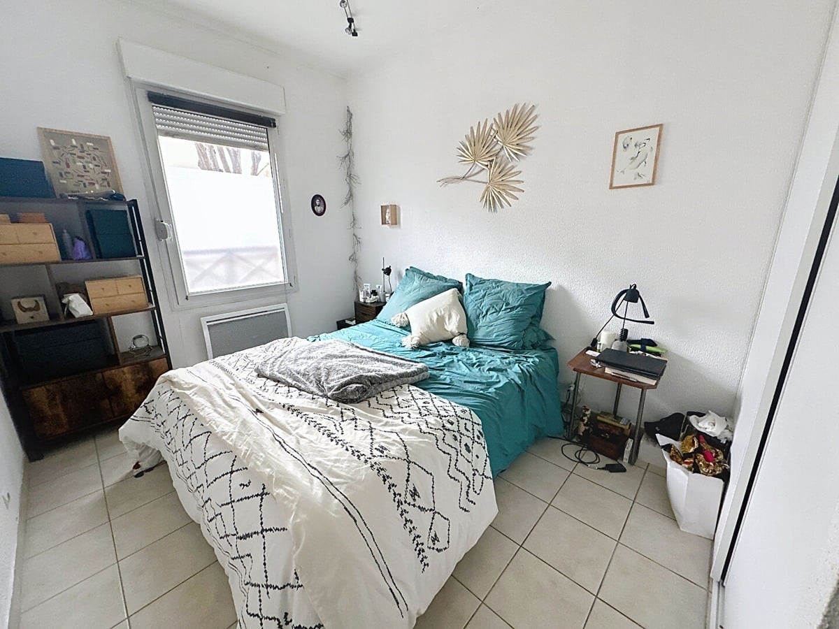Chambre avec lit deux places, couette blanche à motifs géométriques noirs et oreillers turquoise. Étagère métallique noire avec boîtes de rangement, tables de chevet en bois de part et d'autre, décoration murale en feuilles de palmier séchées et fenêtre avec volet roulant.