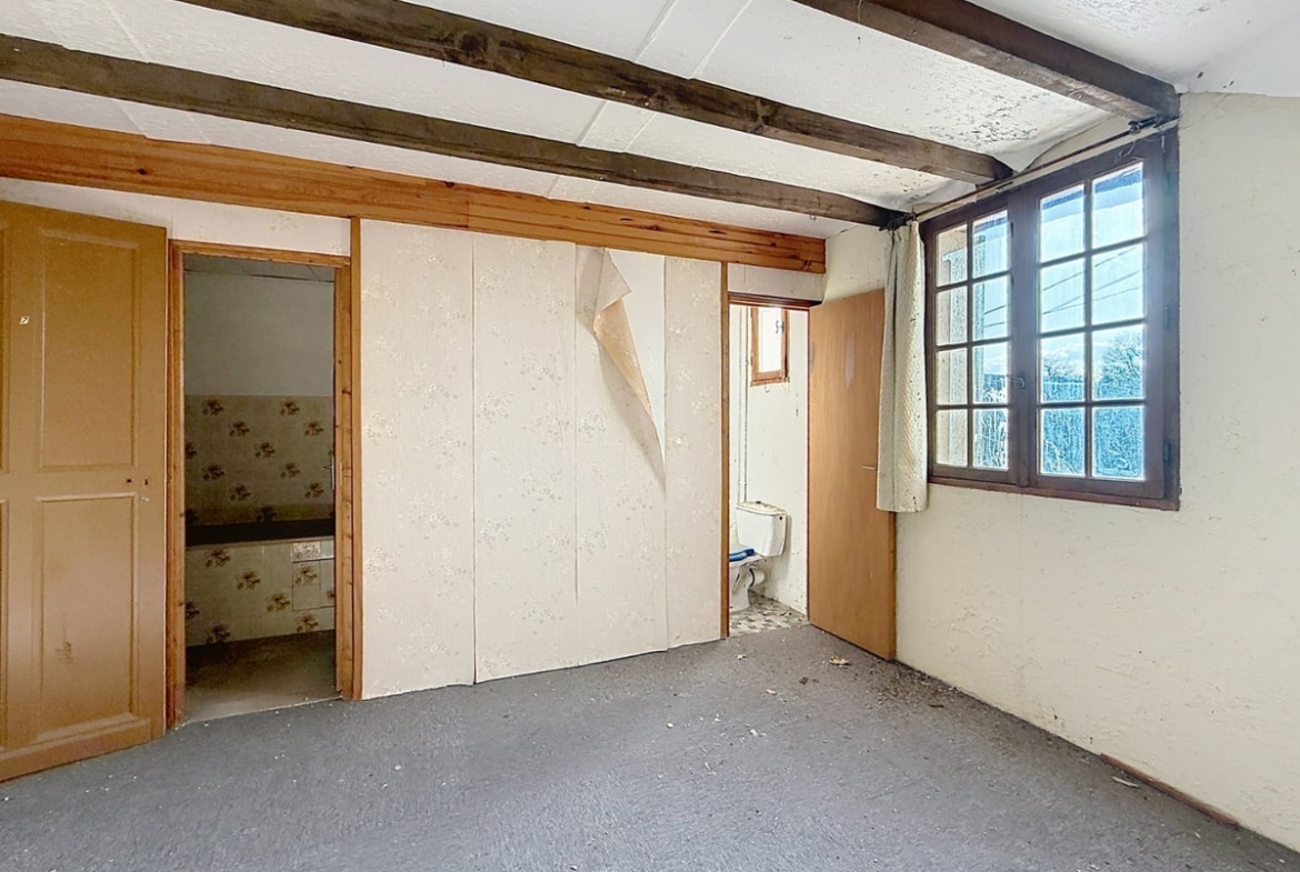 Pièce avec moquette grise usée et murs blancs, équipée de placards intégrés à portes blanches dont le revêtement se décolle. Porte en bois clair ouvrant sur une salle d'eau avec carrelage floral, fenêtre à petits carreaux avec rideau beige et WC visible dans un espace séparé.