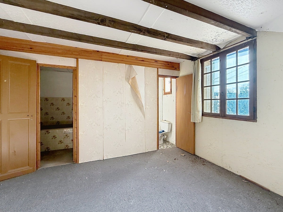 Pièce avec moquette grise usée et murs blancs, équipée de placards intégrés à portes blanches dont le revêtement se décolle. Porte en bois clair ouvrant sur une salle d'eau avec carrelage floral, fenêtre à petits carreaux avec rideau beige et WC visible dans un espace séparé.