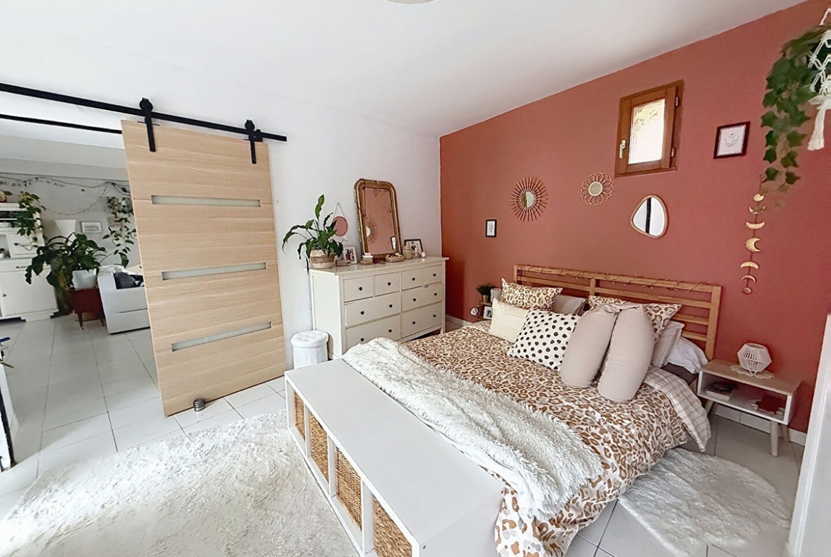 Chambre avec mur d'accent terracotta, lit double avec cadre en bois et linge de lit à motifs léopard. Commode blanche à tiroirs, porte coulissante style grange en bois clair sur rail noir et plantes suspendues.