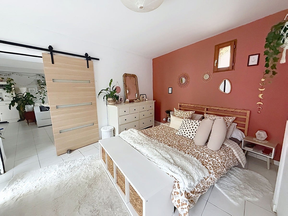 Chambre avec mur d'accent terracotta, lit double avec cadre en bois et linge de lit à motifs léopard. Commode blanche à tiroirs, porte coulissante style grange en bois clair sur rail noir et plantes suspendues.