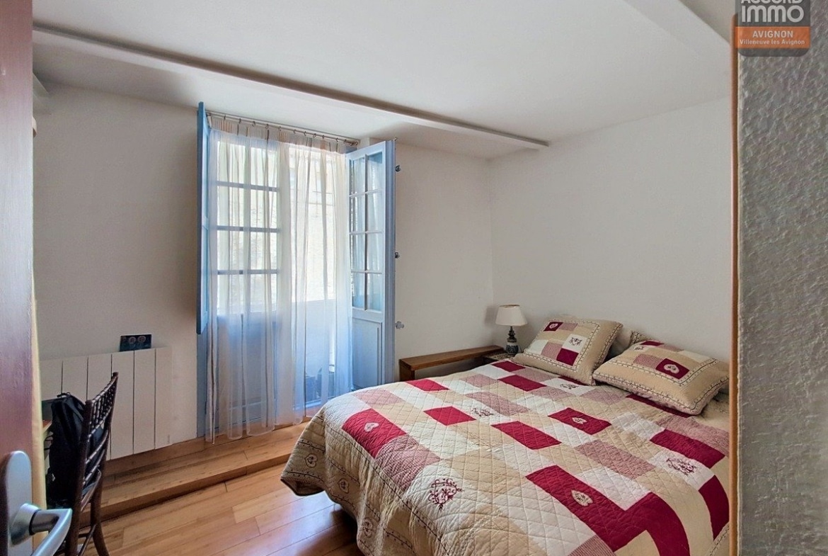 Chambre avec parquet en bois clair et porte-fenêtre à petits carreaux peinte en bleu ouvrant sur balcon. Lit double avec couvre-lit patchwork beige et bordeaux, mur texturé gris et poutre blanche au plafond.