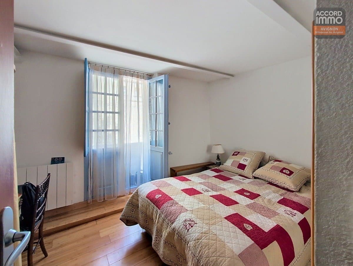 Chambre avec parquet en bois clair et porte-fenêtre à petits carreaux peinte en bleu ouvrant sur balcon. Lit double avec couvre-lit patchwork beige et bordeaux, mur texturé gris et poutre blanche au plafond.