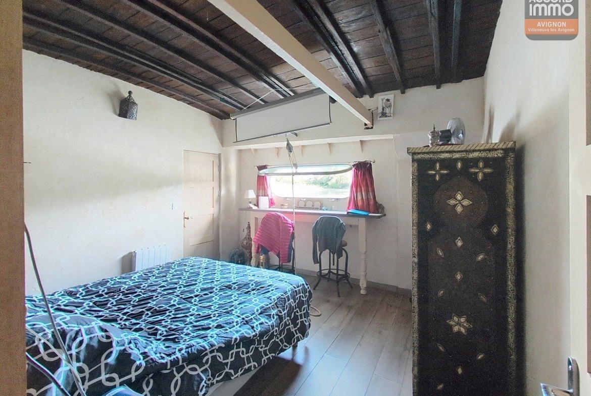 Chambre sous plafond en bois sombre avec poutres apparentes, lit double avec couverture à motifs géométriques bleu et blanc. Armoire décorative orientale noire et dorée, bureau sous fenêtre avec rideaux roses, sol en parquet clair et porte blanche à gauche.