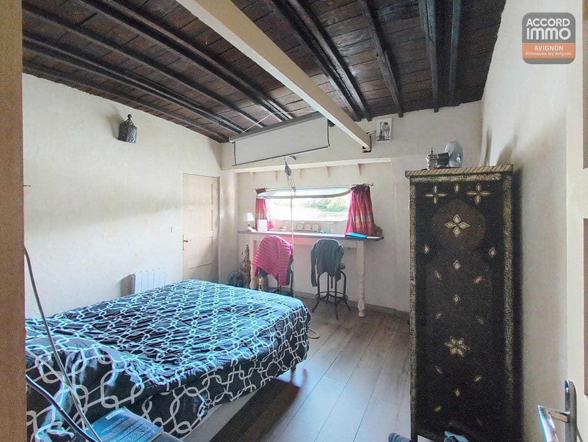 Chambre sous plafond en bois sombre avec poutres apparentes, lit double avec couverture à motifs géométriques bleu et blanc. Armoire décorative orientale noire et dorée, bureau sous fenêtre avec rideaux roses, sol en parquet clair et porte blanche à gauche.