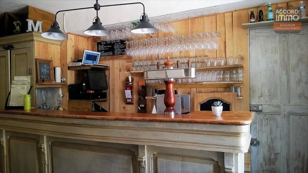 Comptoir de bar patiné blanc avec plateau en bois massif et suspensions industrielles noires. Arrière-bar en pin avec verres suspendus, machine à café professionnelle et caisse enregistreuse.