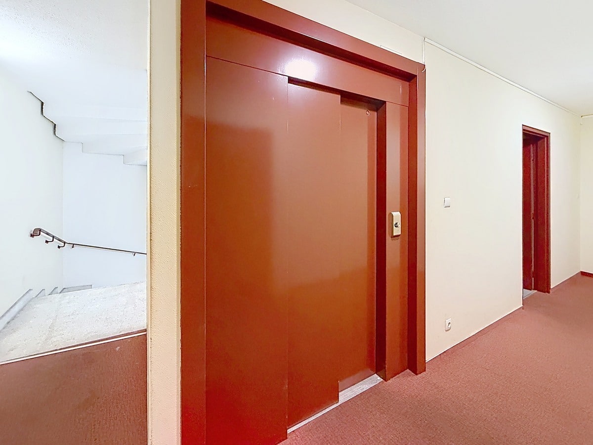 Couloir d'immeuble avec ascenseur aux portes rouge-brun et moquette assortie. Escalier avec rampe métallique visible à gauche, murs blancs et encadrements crème.