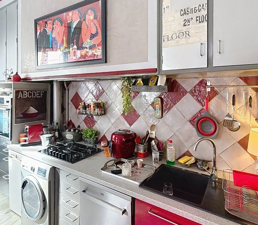 Cuisine équipée avec meubles bas rouges brillants et hauts gris, plan de travail gris moucheté, évier noir. Crédence en carrelage losange rouge et blanc, plaque gaz, lave-linge encastré, four et hotte. Tableau coloré au mur et accessoires de cuisine suspendus sur barre métallique.