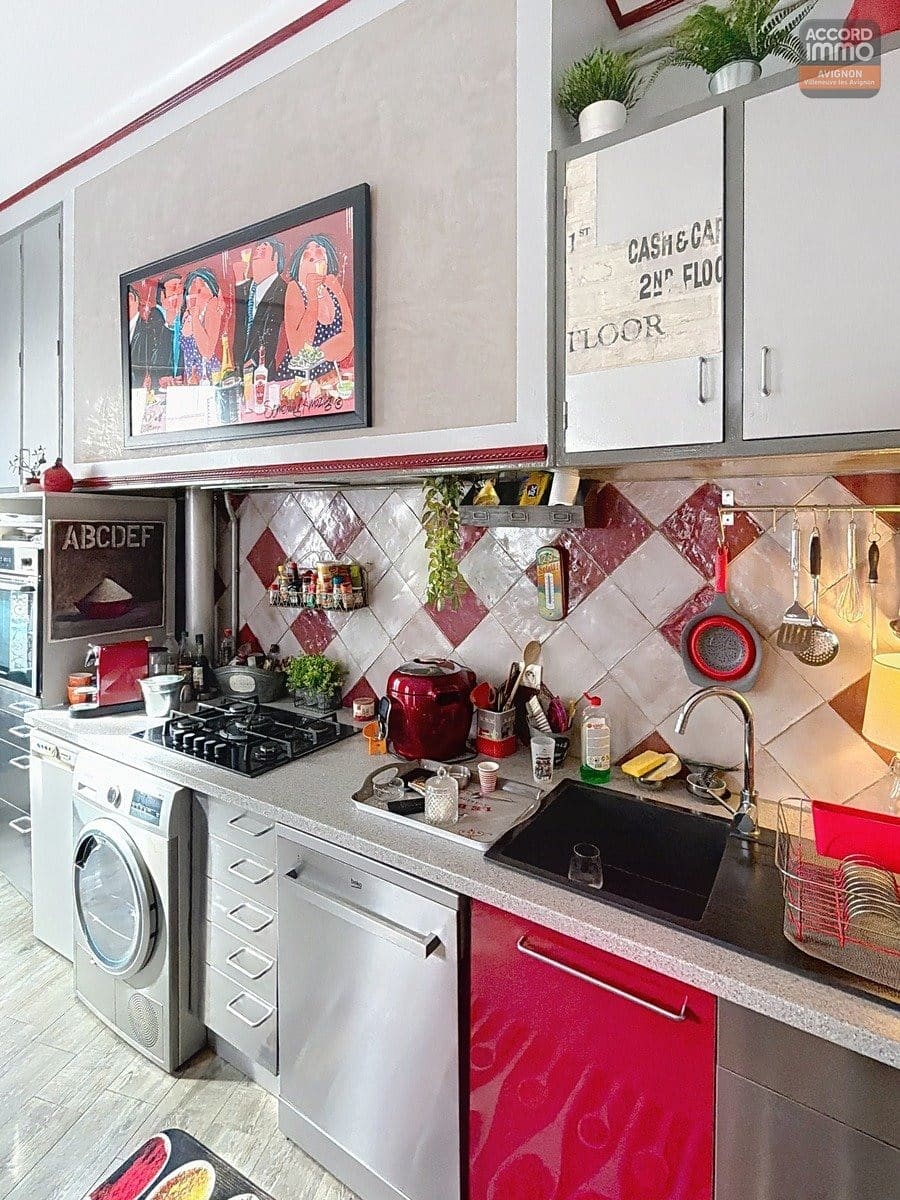 Cuisine équipée avec meubles bas rouges brillants et hauts gris, plan de travail gris moucheté, évier noir. Crédence en carrelage losange rouge et blanc, plaque gaz, lave-linge encastré, four et hotte. Tableau coloré au mur et accessoires de cuisine suspendus sur barre métallique.