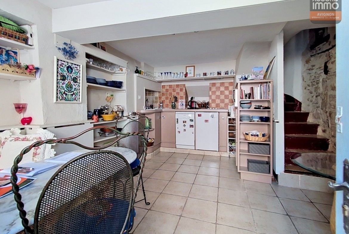 Cuisine ouverte avec sol en carrelage beige, crédence à damier terre cuite et meubles roses. Table ronde en verre avec chaises en fer forgé, étagères ouvertes garnies de vaisselle bleue et escalier en pierre apparente.