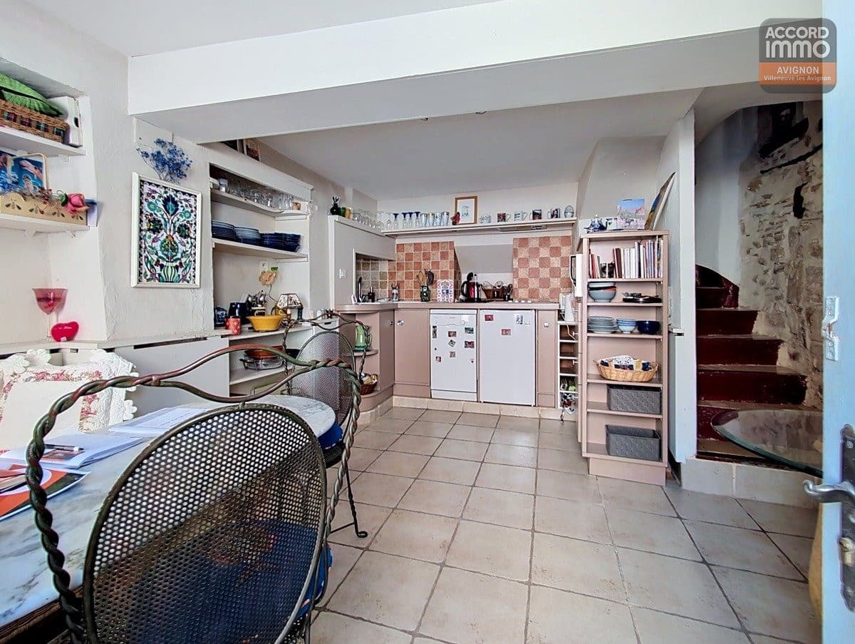 Cuisine ouverte avec sol en carrelage beige, crédence à damier terre cuite et meubles roses. Table ronde en verre avec chaises en fer forgé, étagères ouvertes garnies de vaisselle bleue et escalier en pierre apparente.