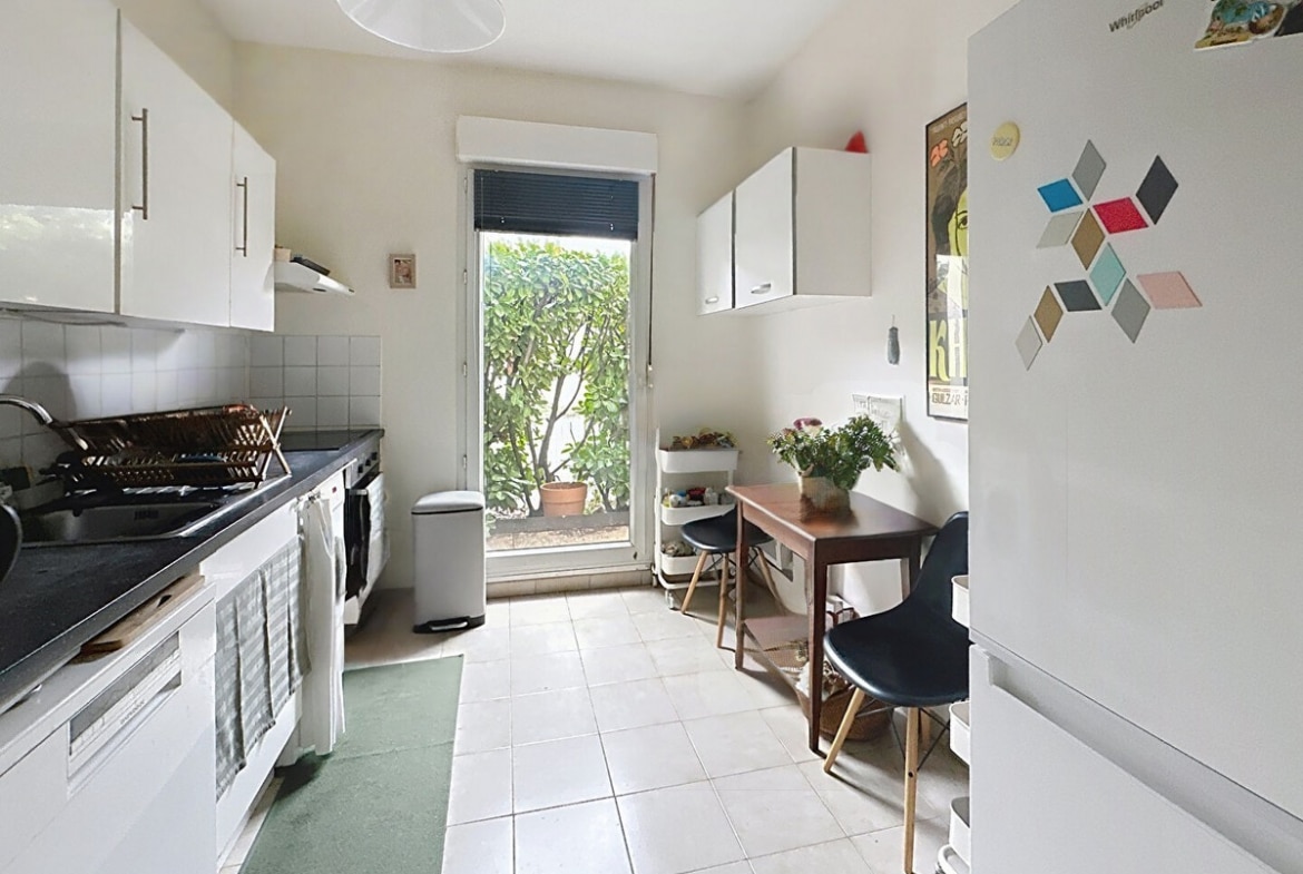 Cuisine en longueur avec meubles blancs, plan de travail noir, égouttoir en osier et porte-fenêtre donnant sur végétation extérieure. Petite table en bois avec deux chaises noires style scandinave, réfrigérateur blanc Whirlpool avec magnets colorés et carrelage beige au sol.