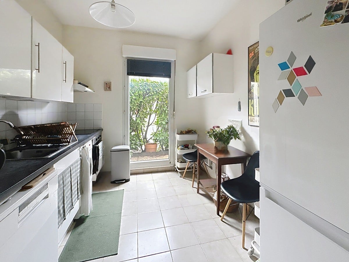 Cuisine en longueur avec meubles blancs, plan de travail noir, égouttoir en osier et porte-fenêtre donnant sur végétation extérieure. Petite table en bois avec deux chaises noires style scandinave, réfrigérateur blanc Whirlpool avec magnets colorés et carrelage beige au sol.