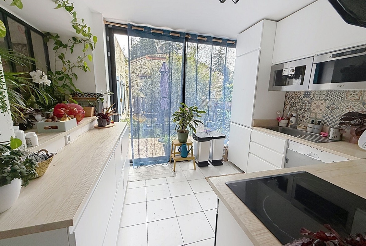 Cuisine aménagée en U avec meubles blancs, plan de travail en bois clair et crédence carreaux de ciment. Baie vitrée coulissante donnant sur jardin, four encastré, plaque induction et nombreuses plantes vertes sur rebord de fenêtre.