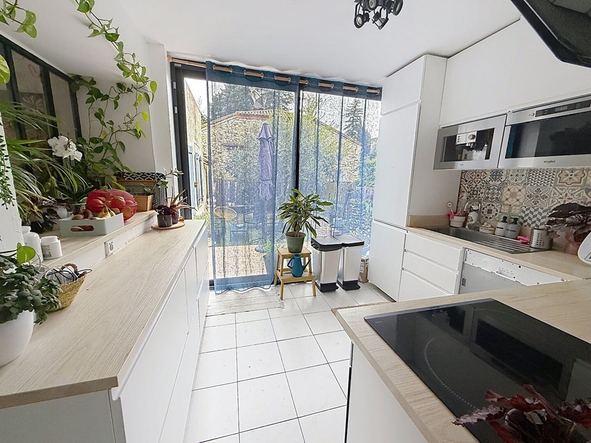 Cuisine aménagée en U avec meubles blancs, plan de travail en bois clair et crédence carreaux de ciment. Baie vitrée coulissante donnant sur jardin, four encastré, plaque induction et nombreuses plantes vertes sur rebord de fenêtre.