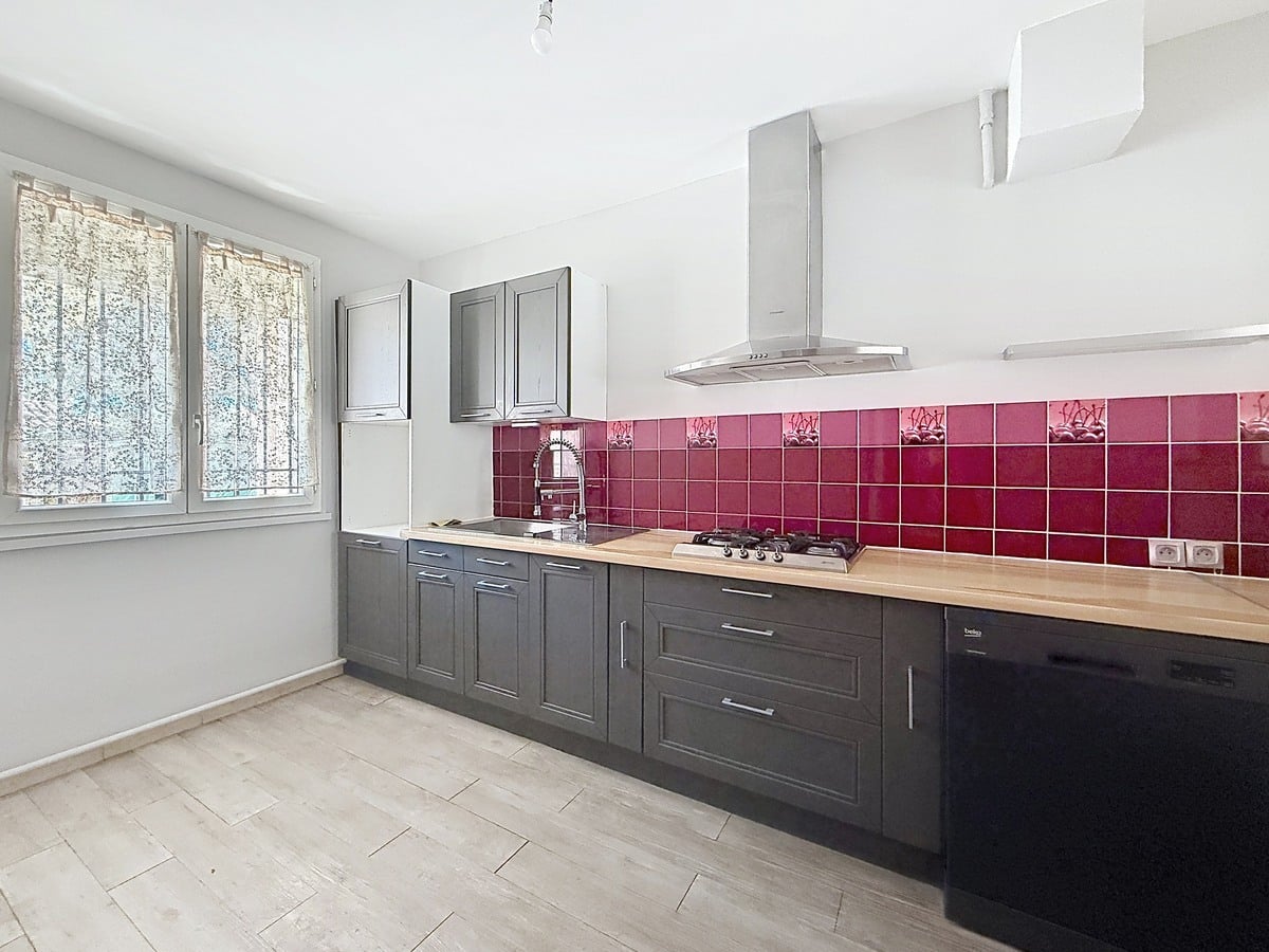 Cuisine linéaire avec façades grises et plan de travail en bois clair. Crédence en carreaux carrés bordeaux et rouge, plaque de cuisson gaz quatre feux, hotte inox murale et lave-vaisselle noir intégré.