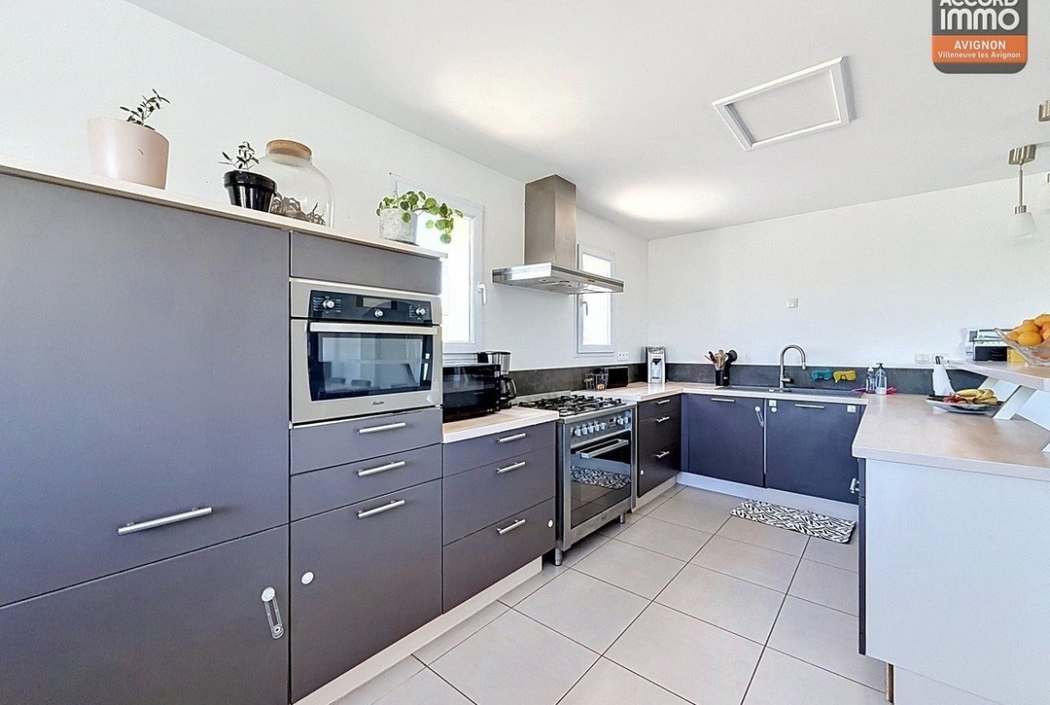 Cuisine en L avec meubles bas et hauts gris anthracite et plan de travail clair. Équipements visibles : four encastré, micro-ondes, hotte en inox et cuisinière avec plaques de cuisson.