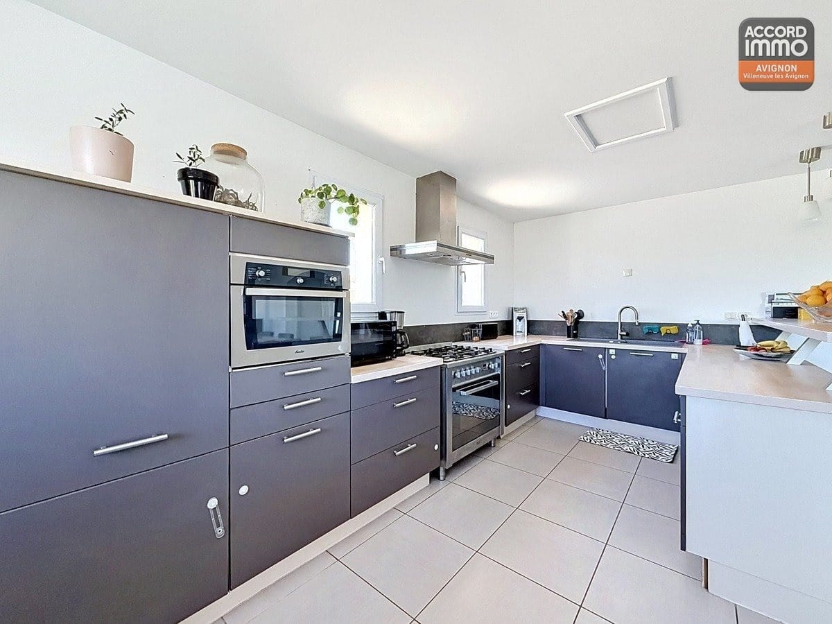 Cuisine en L avec meubles bas et hauts gris anthracite et plan de travail clair. Équipements visibles : four encastré, micro-ondes, hotte en inox et cuisinière avec plaques de cuisson.