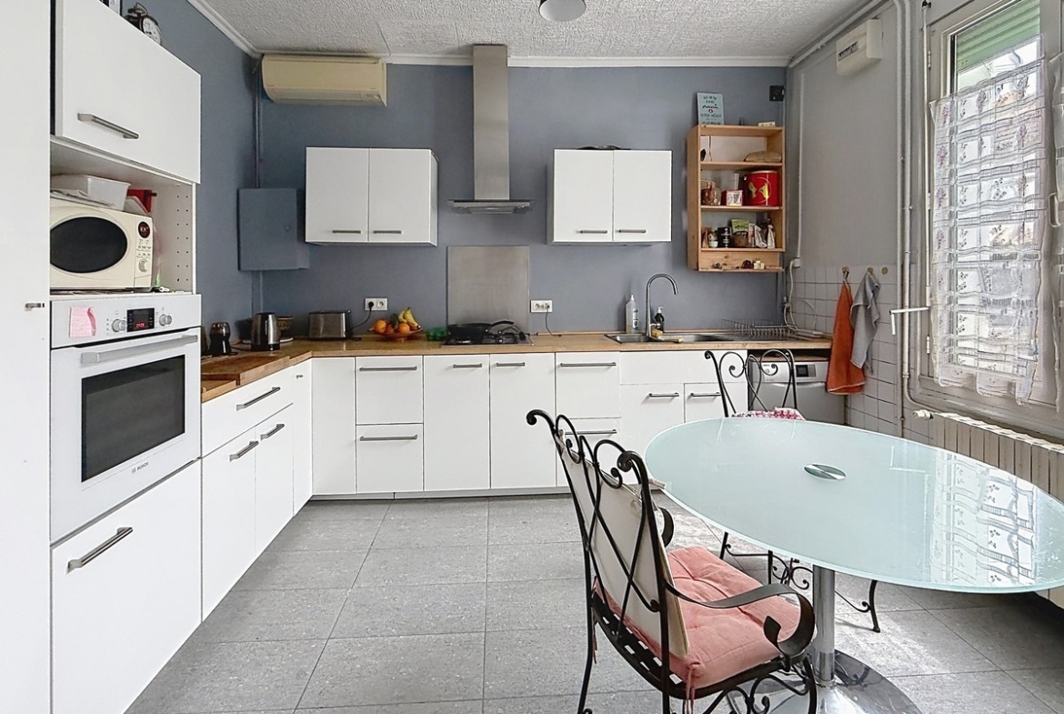 Cuisine en L avec meubles blancs et plan de travail en bois clair. Four encastré, hotte inox et murs peints en bleu-gris. Table ronde en verre avec chaises en fer forgé.