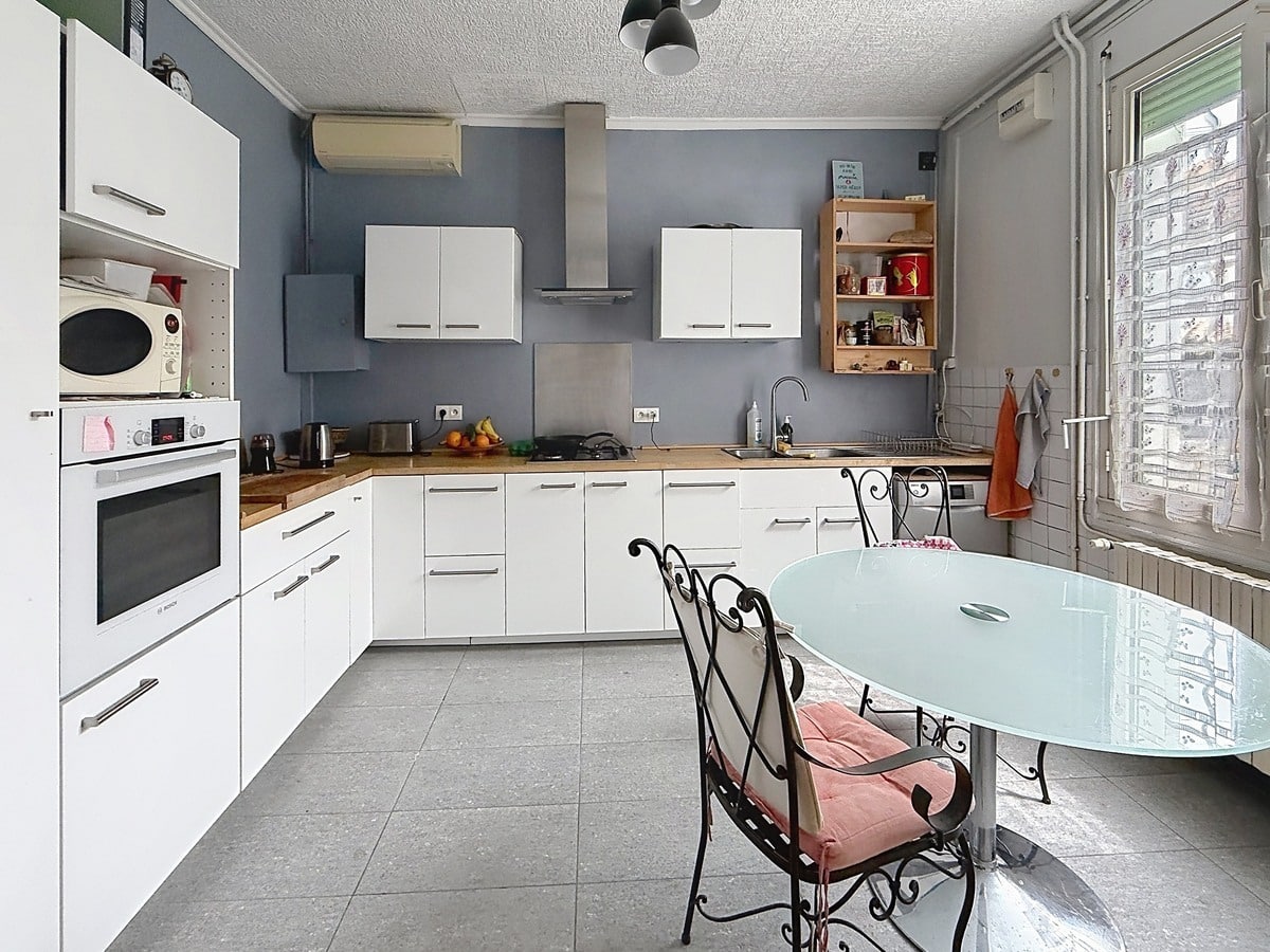 Cuisine en L avec meubles blancs et plan de travail en bois clair. Four encastré, hotte inox et murs peints en bleu-gris. Table ronde en verre avec chaises en fer forgé.
