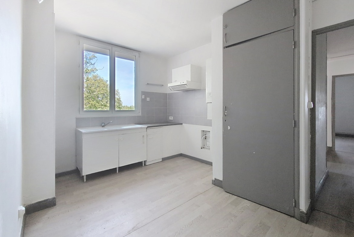 Cuisine avec meubles bas blancs, évier inox et hotte aspirante blanche, crédence carrelée grise. Porte d'entrée métallique grise, fenêtre double vitrage avec vue sur verdure.