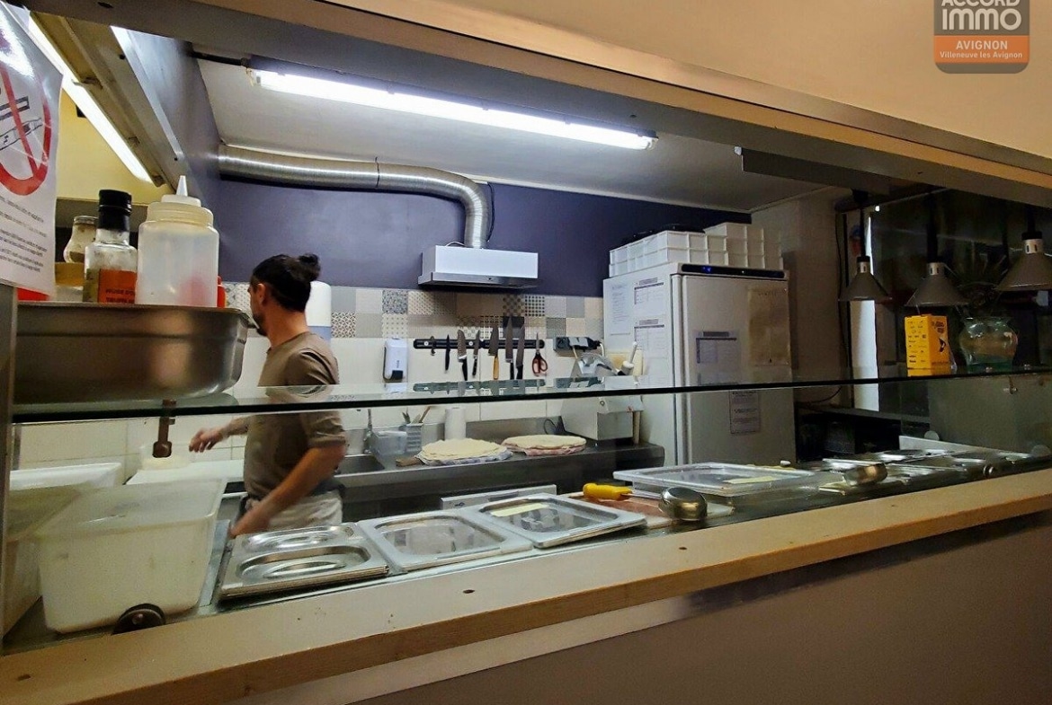 Cuisine ouverte de pizzeria visible depuis le comptoir avec plan de travail en inox et bacs gastronomes. Un cuisinier prépare une pâte à pizza, mur carrelé à motifs, rangée de couteaux magnétique et équipements professionnels en arrière-plan.