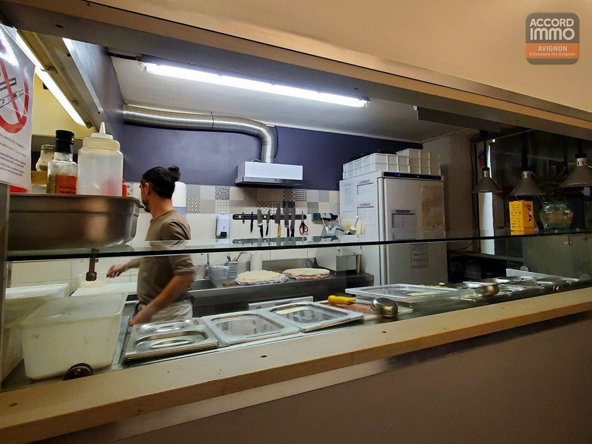 Cuisine ouverte de pizzeria visible depuis le comptoir avec plan de travail en inox et bacs gastronomes. Un cuisinier prépare une pâte à pizza, mur carrelé à motifs, rangée de couteaux magnétique et équipements professionnels en arrière-plan.