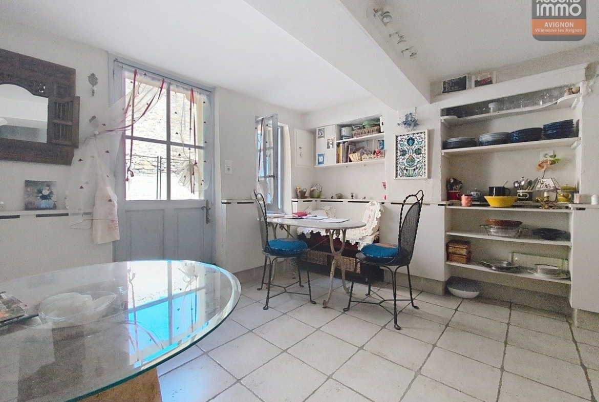 Espace cuisine avec porte d'entrée vitrée peinte en bleu-gris ouvrant sur l'extérieur. Table ronde en marbre, chaises bistrot en fer forgé à assises bleues, étagères murales blanches avec vaisselle et sol carrelé clair.