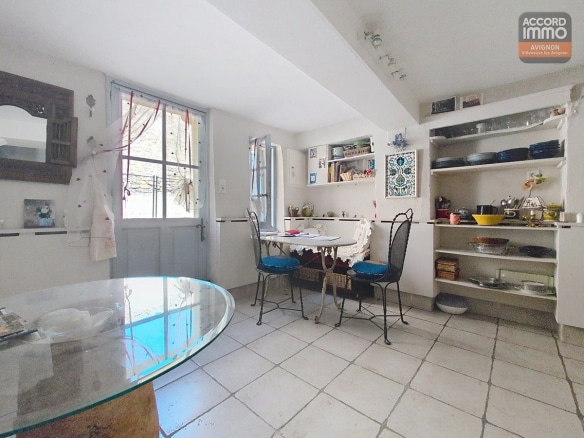 Espace cuisine avec porte d'entrée vitrée peinte en bleu-gris ouvrant sur l'extérieur. Table ronde en marbre, chaises bistrot en fer forgé à assises bleues, étagères murales blanches avec vaisselle et sol carrelé clair.